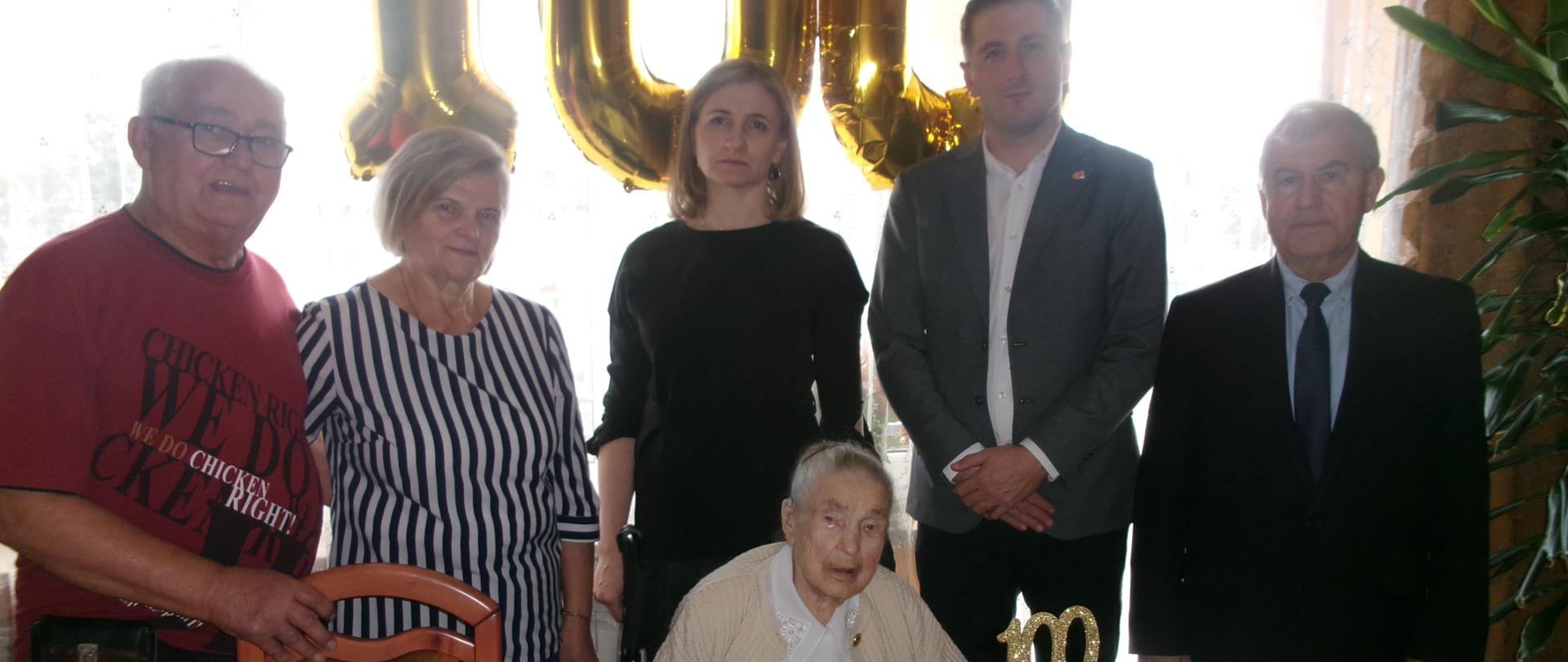 W pomieszczeniu, za stołem, na którym stoi biały tort, siedzi starsza kobieta – jubilatka. Za jej plecami stoi trzech mężczyzn i dwie kobiety. W tle złote balony w kształcie cyfr liczby 100.