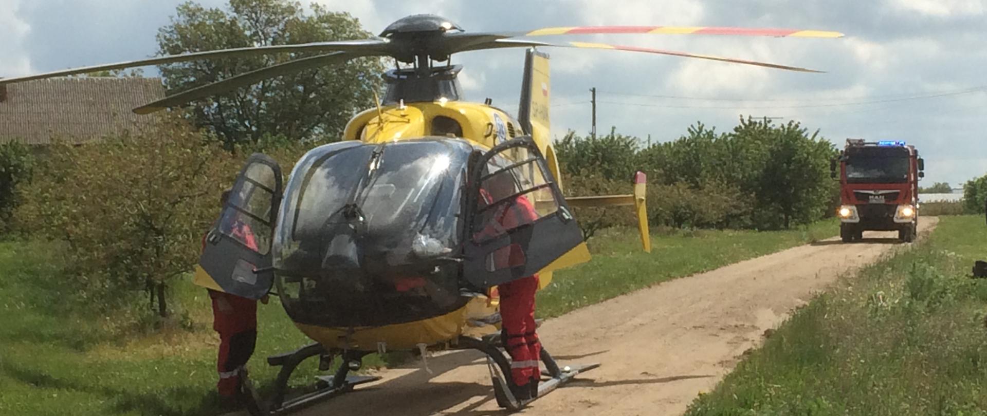 Na zdjęciu na pierwszym [planie stojący na polnej drodze śmigłowiec LPR oraz znajdujących się przy jago otwartych drzwiach, po obu stronach, ratowników. Z tyłu widać stojący samochód strażacki.