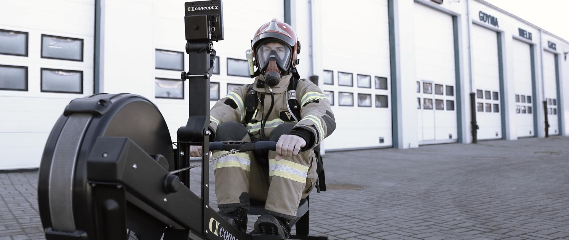 W tle garaże. Strażak ubrany w nomex, hełm, maskę twarzową korzysta z ergonometru.