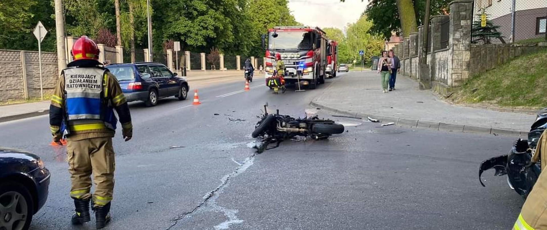 Strażak kierujący działaniami, w środku przewrócony motocykl , w tle zastępy JRG