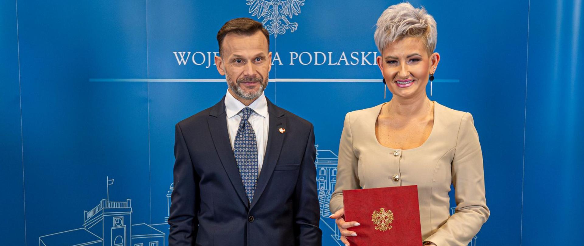 Mężczyzna w garniturze stoi obok kobiety, która w ręku trzyma dokument z godłem Polski. W tle baner z napisem Wojewoda Podlaski.