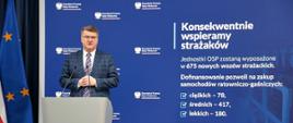 Maciej Wąsik przemawia przy mównicy w trakcie konferencji prasowej za nim banner mswia i napisami konsekwentnie wspieramy strażaków. Jednostki OSP zostaną wyposażone w 675 nowych wozów strażackich. dofinansowanie pozwoli na zakup samochodów ratowniczo-gaśniczych ciężkich 78 średnich 417 lekkich 180.