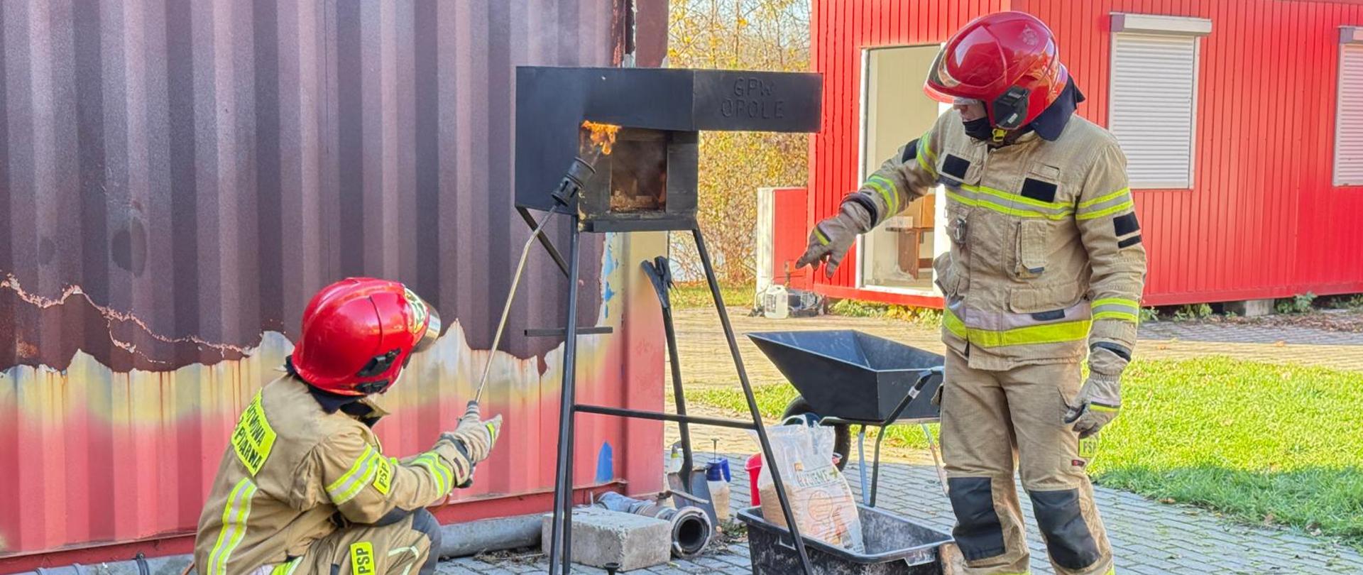 Dwóch strażaków pracuje przy metalowym stanowisku z otwartym płomieniem, analizując jego zachowanie. Obok znajduje się wózek z narzędziami i substancjami pomocniczymi, co sugeruje ćwiczenia z zakresu rozpoznawania i gaszenia pożarów różnych materiałów. Tło z nadpaloną ścianą budynku świadczy o intensywności wcześniejszych ćwiczeń.