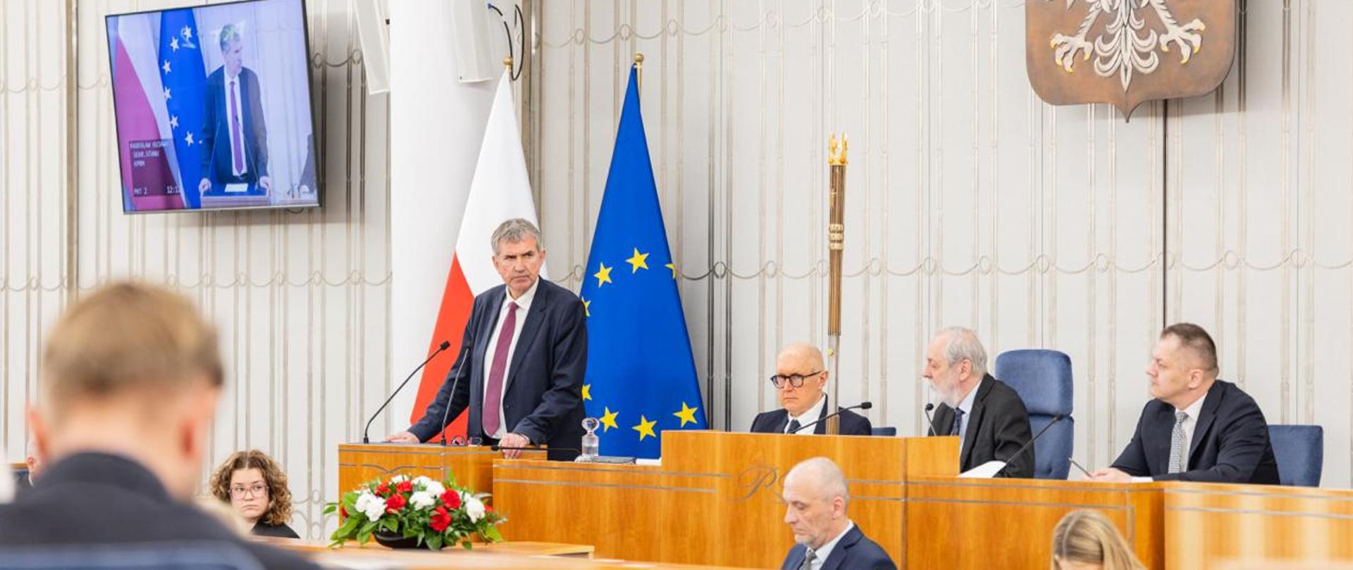 Zdjęcie przedstawia salę posiedzeń Senatu. Za mównicą Radosław Kujawa, sekretarz stanu w KPRM. Za mównicą stoją dwie flagi - po lewej polska (biało-czerwona), natomiast po prawej flaga UE (niebieska z żółtymi gwiazdkami). Po prawej stronie za stołem prezydialnym siedzi trzech mężczyzn - w środku wicemarszałek Senatu Rafał Grupiński, który przewodniczy obradom. Obok wicemarszałka znajduje się laska marszałkowska. Nad stołem prezydialnym po prawej stronie zdjęcia wisi godło Polski (orzeł). Poniżej stołu prezydialnego w tzw. "fosie" siedzi trzech urzędników Kancelarii Senatu (dwie kobiety i jeden mężczyzna), którzy odpowiadają za obsługę obrad. Na stole urzędników znajduje się biało-czerwona kompozycja kwiatowa. Po lewej stronie zdjęcia (u góry) znajduje się monitor z przekazem na żywo z obrad Senatu (wyświetlony wizerunek ministra R. Kujawy). W lewym dolnym rogu znajduje się rozmazany wizerunek siedzącej tyłem osoby.