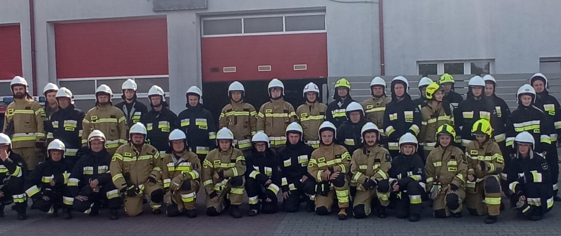 Na zdjęciu widoczni są druhowie OSP ustawieni do wspólnego zdjęcia. Ubrani są w ubrania specjalne i hełmy. W tle widać bramy garażowe Jednostki Ratowniczo-Gaśniczej Nr 2 w Koninie.
