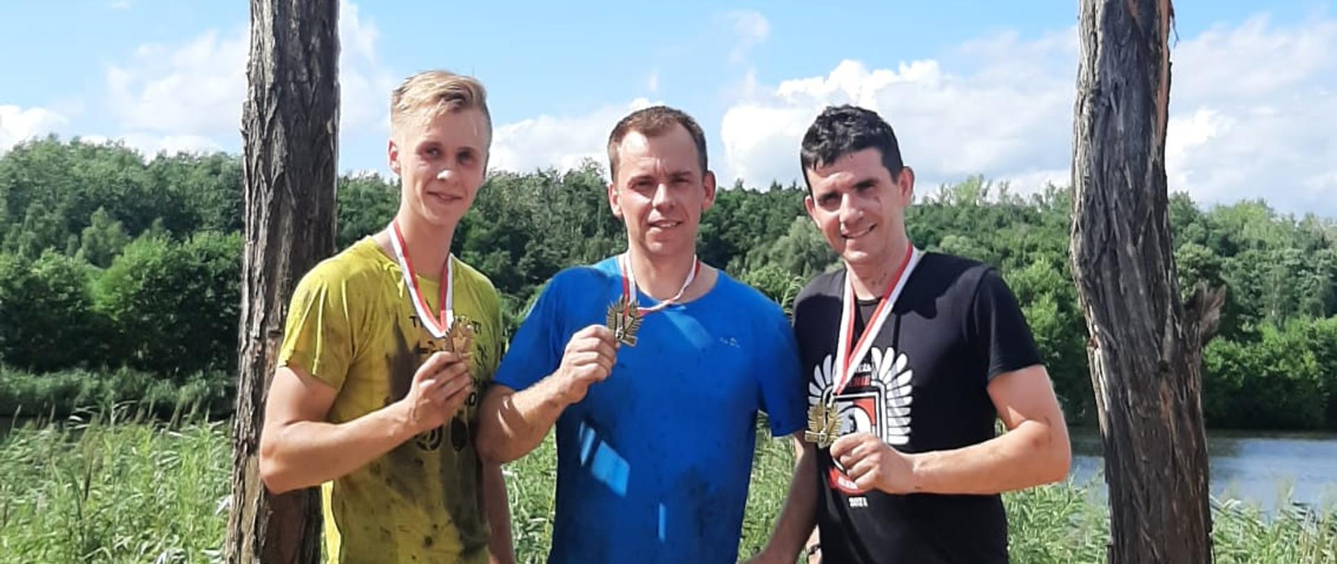 Na zdjęciu widzimy trzech mężczyzn stojących w rzędzie, którzy w dłoniach trzymają medale. Mężczyzna z lewej bardzo ubrudzony ubrane jest w koszulkę koloru żółtego oraz czarne spodenki. Mężczyzna po środku również mocno zabrudzony ubrany jest w koszulkę koloru niebieskiego oraz czarne spodenki. Mężczyzna po prawej ubrany w czarną koszulkę oraz niebieski spodenki. Mężczyźni po wyrazach twarzy zmęczenie ale zadowoleni i uśmiechnięci. Mężczyźni stoją pomiędzy drewnianymi słupami do których przymocowana jest decha z czarnym napisem " Borowy staw". Dalej w kadrze widzimy zarośla, drzewa oraz zbiornik wodny. 