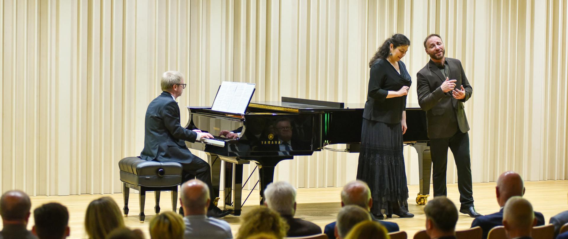 Na scenie sali koncertowej odbywa się występ muzyczny z udziałem trojga artystów. Pianista siedzi przy czarnym fortepianie i akompaniuje dwójce śpiewaków – kobiecie w długiej czarnej sukni oraz mężczyźnie w ciemnym garniturze. Artyści występują przed licznie zgromadzoną publicznością. Jasne, pionowe dekoracje ścienne i eleganckie wnętrze podkreślają uroczysty charakter wydarzenia.
