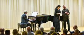 Na scenie sali koncertowej odbywa się występ muzyczny z udziałem trojga artystów. Pianista siedzi przy czarnym fortepianie i akompaniuje dwójce śpiewaków – kobiecie w długiej czarnej sukni oraz mężczyźnie w ciemnym garniturze. Artyści występują przed licznie zgromadzoną publicznością. Jasne, pionowe dekoracje ścienne i eleganckie wnętrze podkreślają uroczysty charakter wydarzenia.