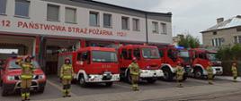 Zdjęcie przedstawia 6 strażaków, stojących w pozycji "na baczność" przed pojazdami pożarniczymi. W tle znajduje się budynek strażnicy.