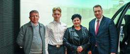 Minister Czarnek, młody mężczyzna w białej koszuli, kobieta w czarnym ubraniu i mężczyzna w szarym garniturze stoją w rzędzie, obok nich połowa przeciętego w poprzek samochodu.