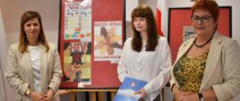 Trzy osoby stoją w pomieszczeniu, na tle sztalug z oprawionymi plakatami edukacyjnymi dotyczącymi profilaktyki HIV/AIDS. Osoba pośrodku trzyma niebieską teczkę oraz torbę z upominkami. Po lewej i prawej stronie stoją osoby ubrane w jasne marynarki.