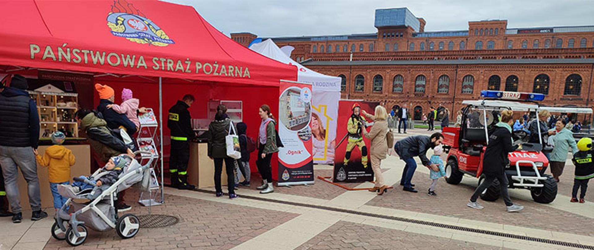 namiot Państwowej Straży Pożarnej na dziedzińcu rynku Manufaktury, widoczni ludzie oglądający wystawkę, obok namiotu kartonowa postać strażaka przy której ludzie robią sobie zdjęcia, stoi samochód elektryczny Melex z napisem straż oraz z lampami błyskowymi na dachu, dzieci przebrane w strażaków wsiadają do samochodu elektrycznego, w tle budynek z czerwonej cegły
