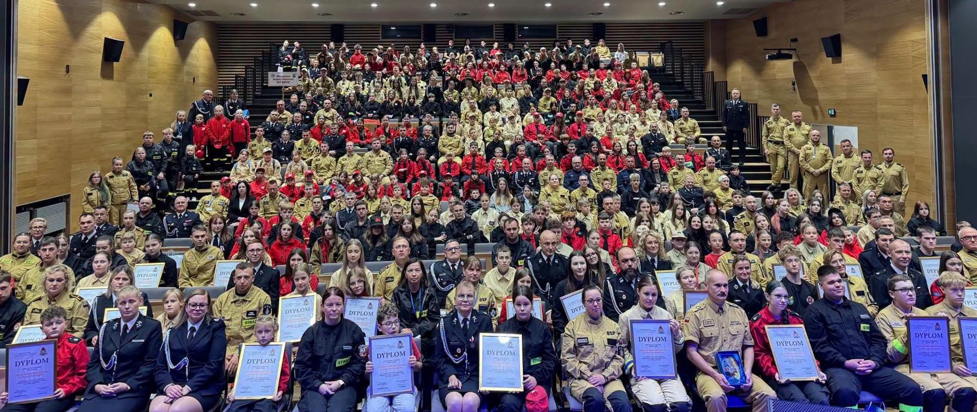 Na fotografii widoczna jest bardzo liczna grupa uczestników III Kongresu zgromadzona w dużej sali widowiskowej. Wśród nich znajdują się strażacy PSP i OSP, członkowie Młodzieżowych Drużyn Pożarniczych oraz zaproszeni goście. Część osób w pierwszych rzędach trzyma dyplomy i wyróżnienia, a zdjęcie ma charakter pamiątkowy.