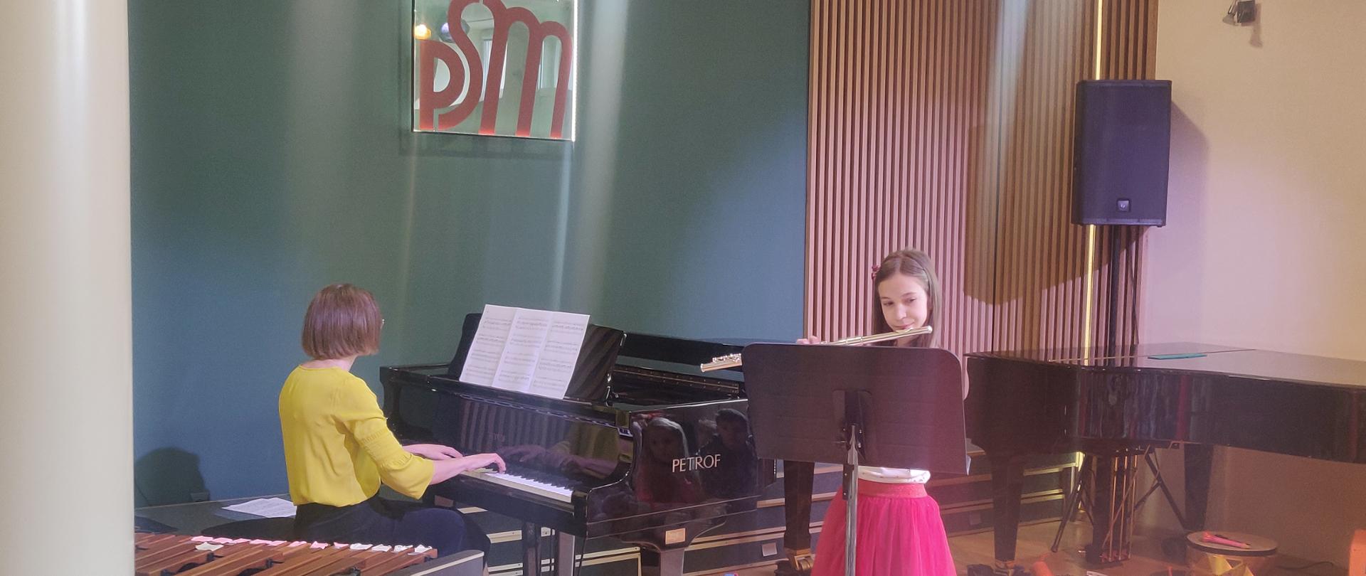 Na zdjęciu widać dziewczynkę w różowej sukience która gra na flecie przed nią stoi pulpit. Po lewej stronie siedzi kobieta przy fortepianie i akompaniuje dziewczynce. Na podłodze po prawej stronie widoczne instrumenty perkusyjne a w tle widoczna ściana w kolorze zielonym z drewnianymi lamelami.