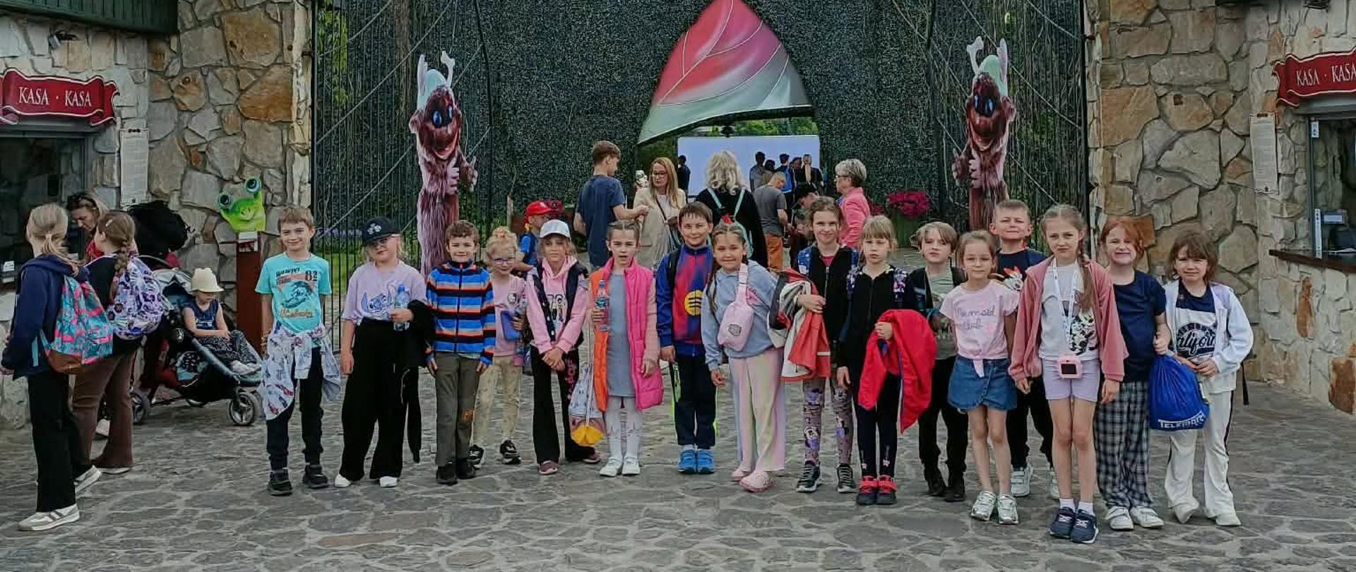 Zdjęcie grupowe kilkanaściorga dzieci stojących przed wejściem do parku tematycznego „Magiczne Ogrody”. Dzieci są ubrane w kolorowe, codzienne ubrania i stoją w rzędzie na kamiennym, brukowanym dziedzińcu. Nad nimi wznosi się wysoki łuk z jasnego kamienia, pod którym wisi duży, zielony szyld ze złotym, ozdobnym napisem „MAGICZNE OGRODY”. W tle, za otwartą bramą, widać unikalną, ciemnozieloną konstrukcję przypominającą wysoki żywopłot oraz kolorowe wejście w kształcie pąka kwiatu. Po lewej i prawej stronie kadru widoczne są fragmenty kamiennych budek z czerwonymi tabliczkami z napisem „KASA”.