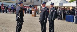 Trzej strażacy w mundurach galowych stoją na środku placu apelowego. Więcej strażaków stoi w prawej części zdjęcia pod namiotem. Na wprost kamienice i przed nimi sztandary. 