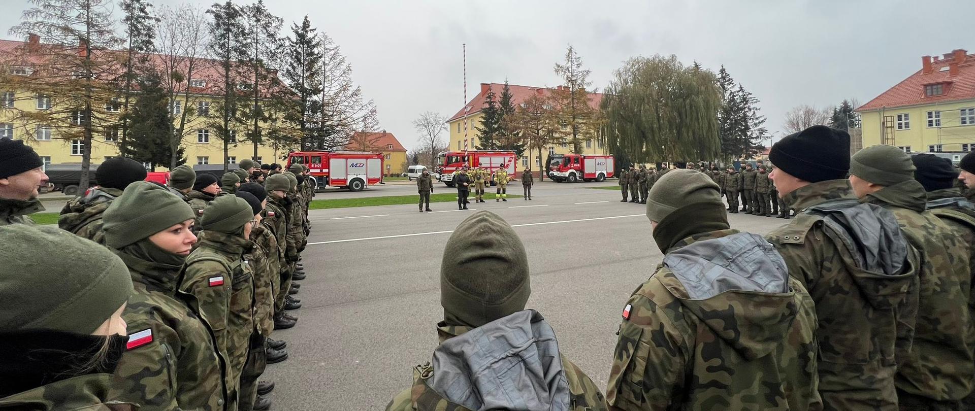 Plac wewnętrzny jednostki wojskowej. Z prawej i lewej strony oraz na dole zdjęcia stoją żołnierze w mundurach zielonych. W oddali trzy samochody strażackie oraz budynki.