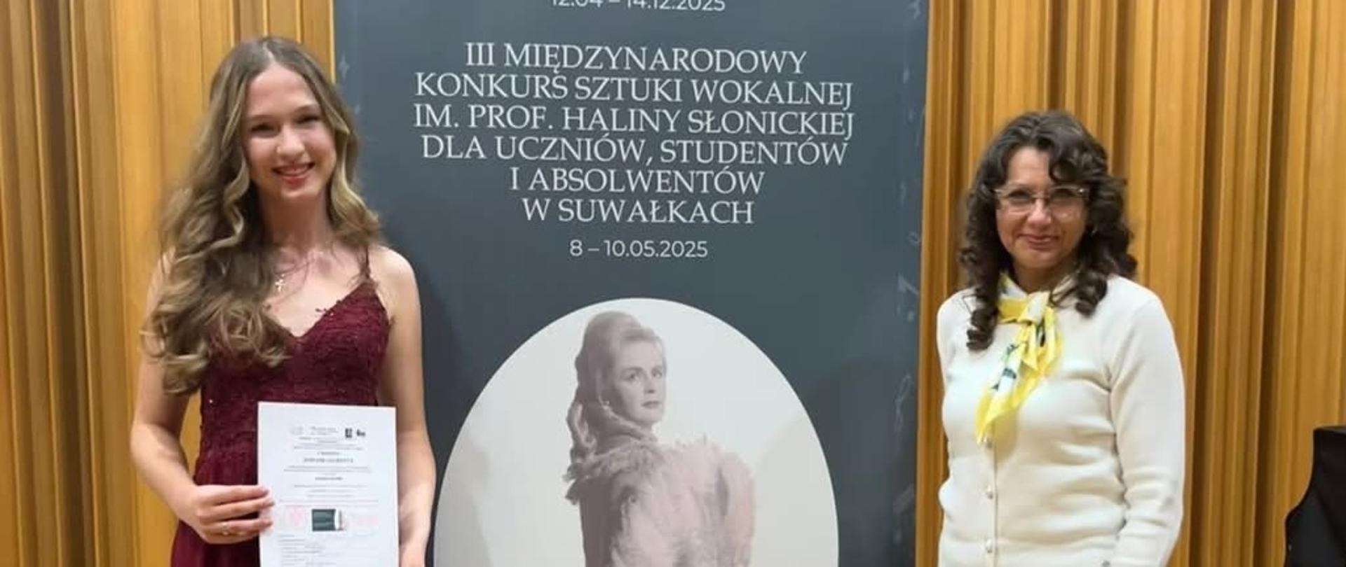 Amelia Szmidt i Monika Brewczak - III Międzynarodowy Konkurs Sztuki Wokalnej w Suwałkach. Uczennica i nauczyciel stojące przy banerze reklamowym konkursu na scenie sali koncertowej.