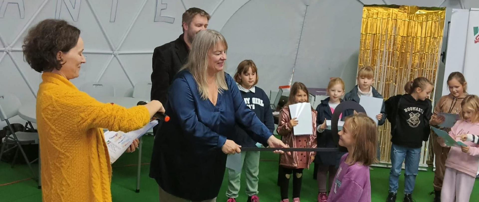 Ceremonia pasowania - dyrektor szkoły pasuje uczennicę dużą czarną półnutą. Obok stoi nauczycielka w żółtym swetrze, nauczyciel i grupa dzieci