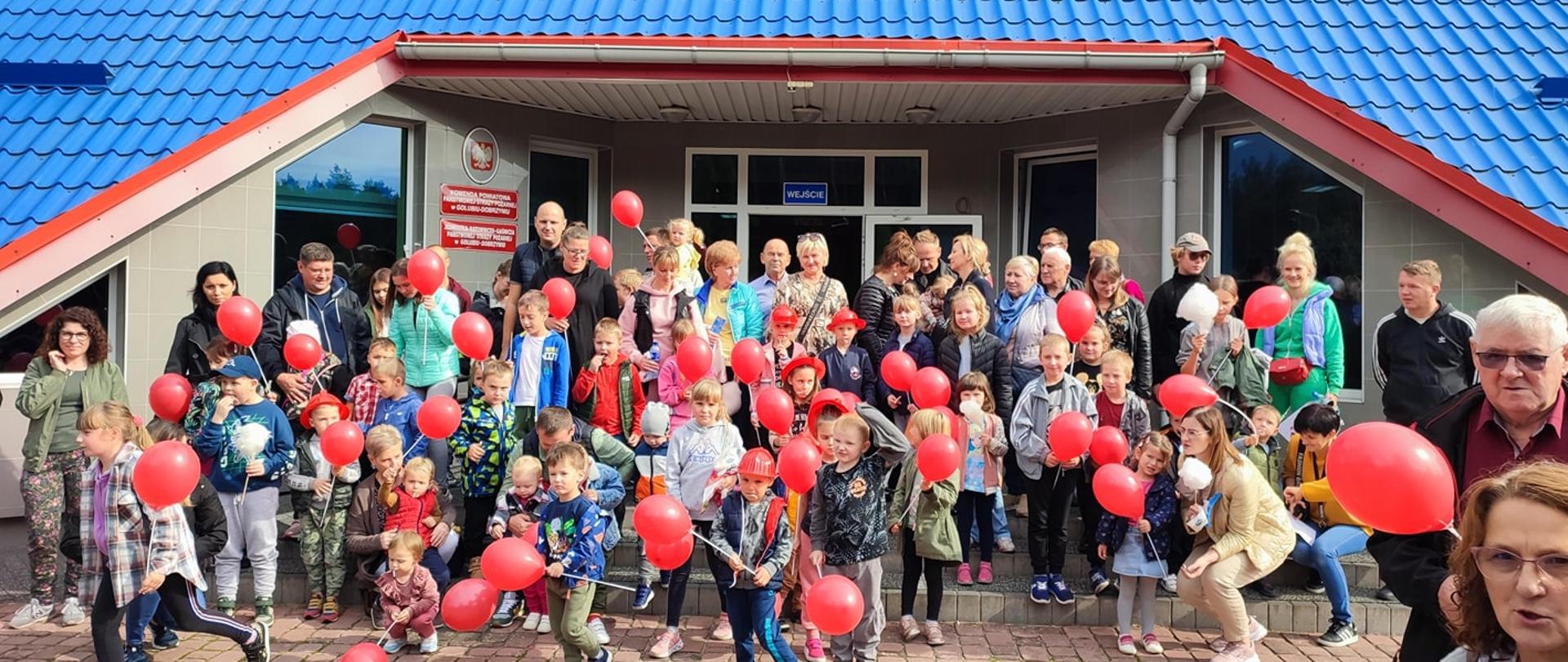 Grupa dzieci i dorosłych stoi na schodach przed budynkiem Komendy Powiatowej PSP w Golubiu-Dobrzyniu. Dzieci trzymają w dłoniach czerwone baloniki, część ma na głowie czerwone hełmy.