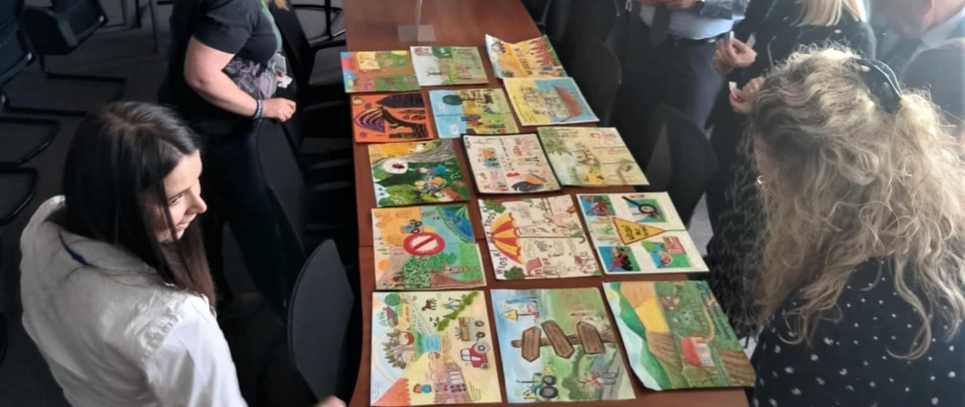 Grupa osób zgromadzonych wokół dużego, drewnianego stołu w sali konferencyjnej ogląda rozłożone na stole kolorowe prace plastyczne, przedstawiające m.in. traktory, dzieci, zwierzęta i krajobrazy wiejskie. W tle widoczna jest ściana z ramkami i zdjęciami, doniczka z dużą rośliną oraz witryna z pucharami. 