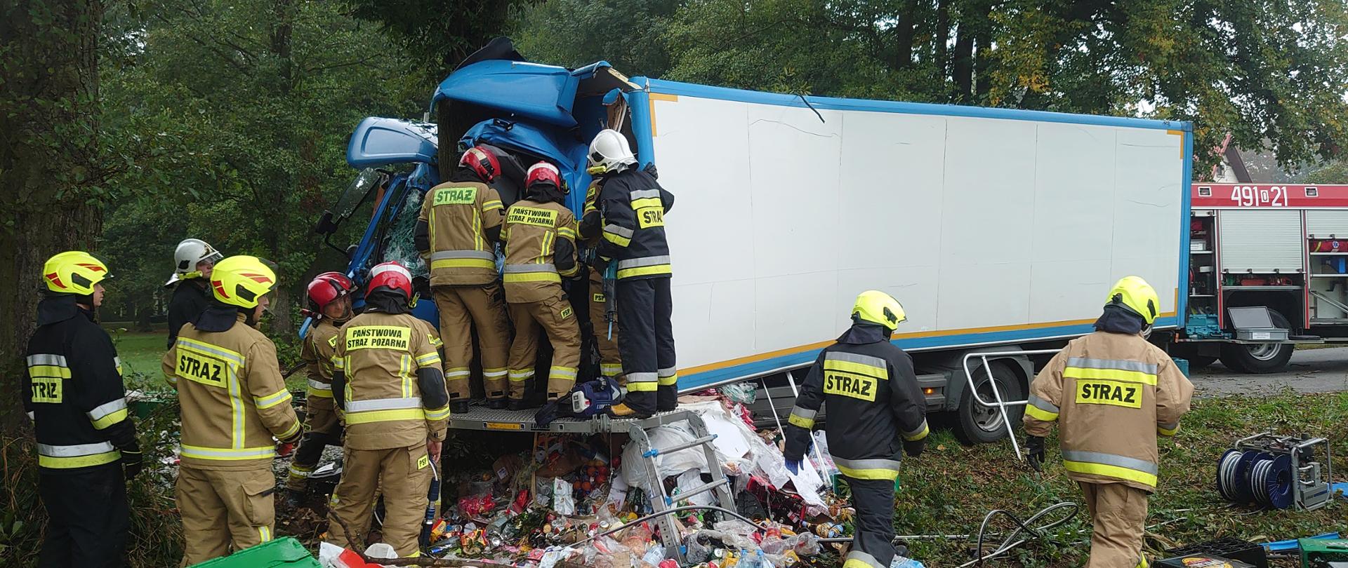 Zdjęcie przedstawia samochód ciężarowy, który uderzył w drzewo oraz strażaków działających przy wraku pojazdu