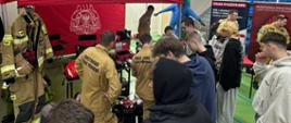 Młodzież przy stoisku PSP ogląda sprzęt i rozmawia ze strażakami o służbie oraz wyposażeniu.