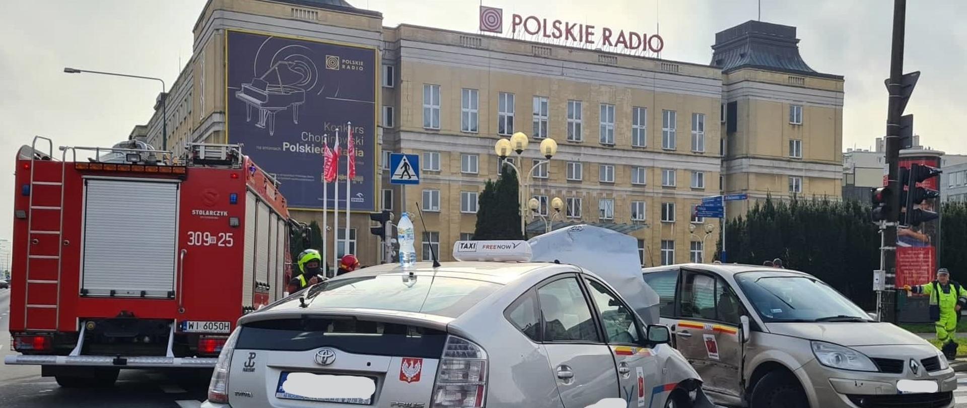 Na zdjęciu widoczne są dwa pojazdy które brały udział w wypadku, oraz pracujący strażacy, w tle samochody straży pożarnej.
