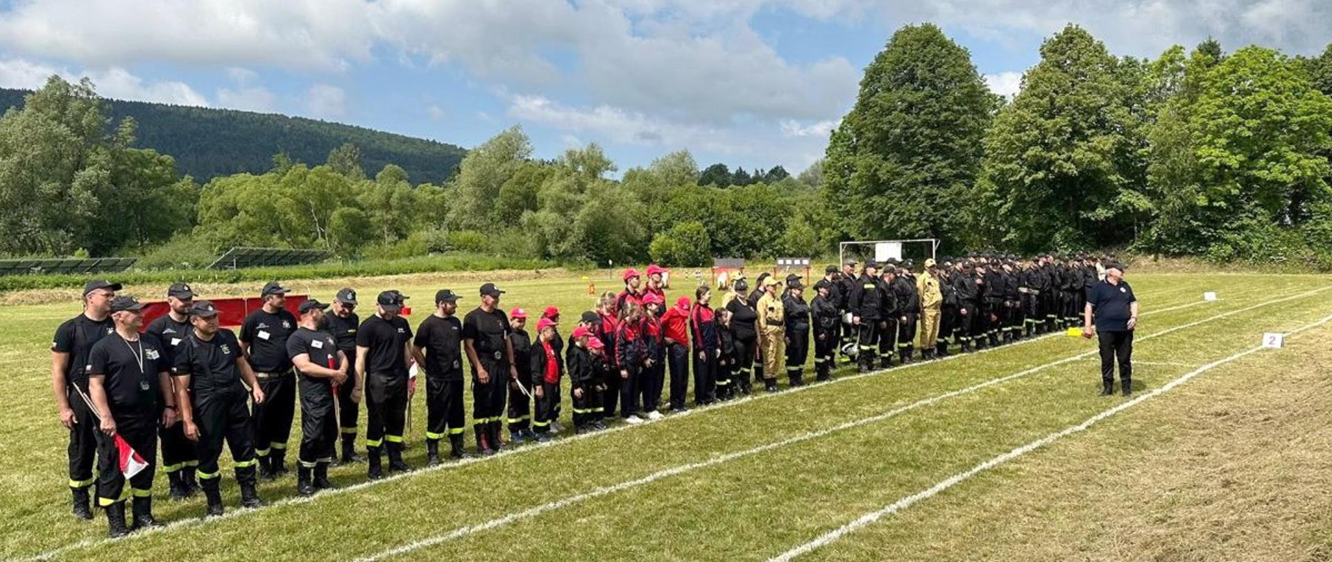 Zdjęcie przedstawia stojące na stadionie drużyny i sędziów biorących udział w zawodach sportowo-pożarniczych. 