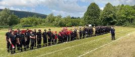 Zdjęcie przedstawia stojące na stadionie drużyny i sędziów biorących udział w zawodach sportowo-pożarniczych. 