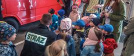 Zdjęcie przedstawia grupę dzieci podczas zapoznania ze sprzętem strażackim w pomieszczeniu garażu na tle pojazdów strażackich koloru czerwonego.