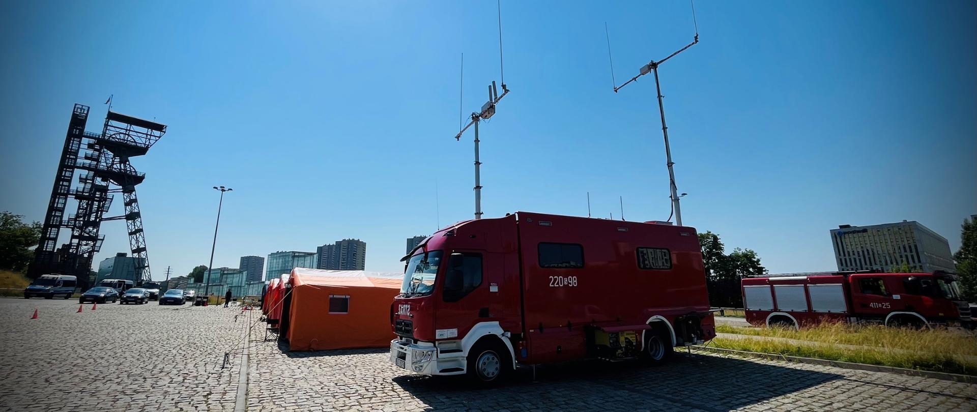 Na zdjęciu widoczny parking, na którym po prawej stronie stoi specjalny samochód dowodzenia i łączności PSP, za nim pomarańczowe namioty. Po lewej stronie zabytkowy szyb kopalni. 