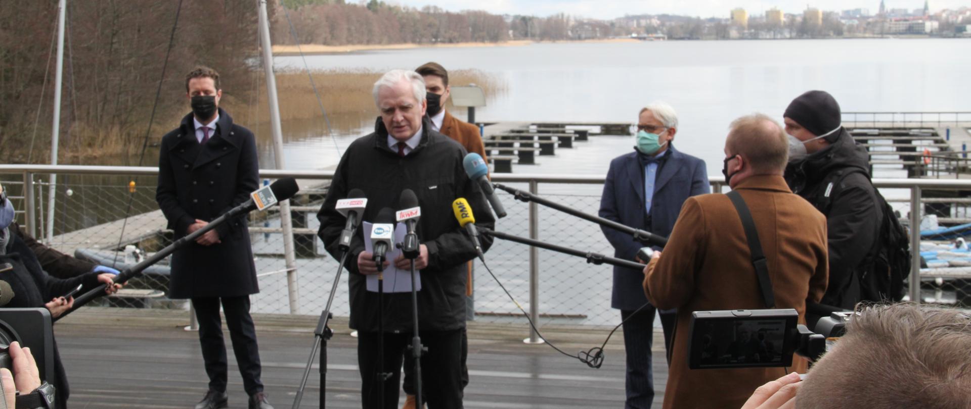Jarosław Gowin podczas konferencji prasowej. Stoi na pomoście, za nim jezioro, przed nim dziennikarze.