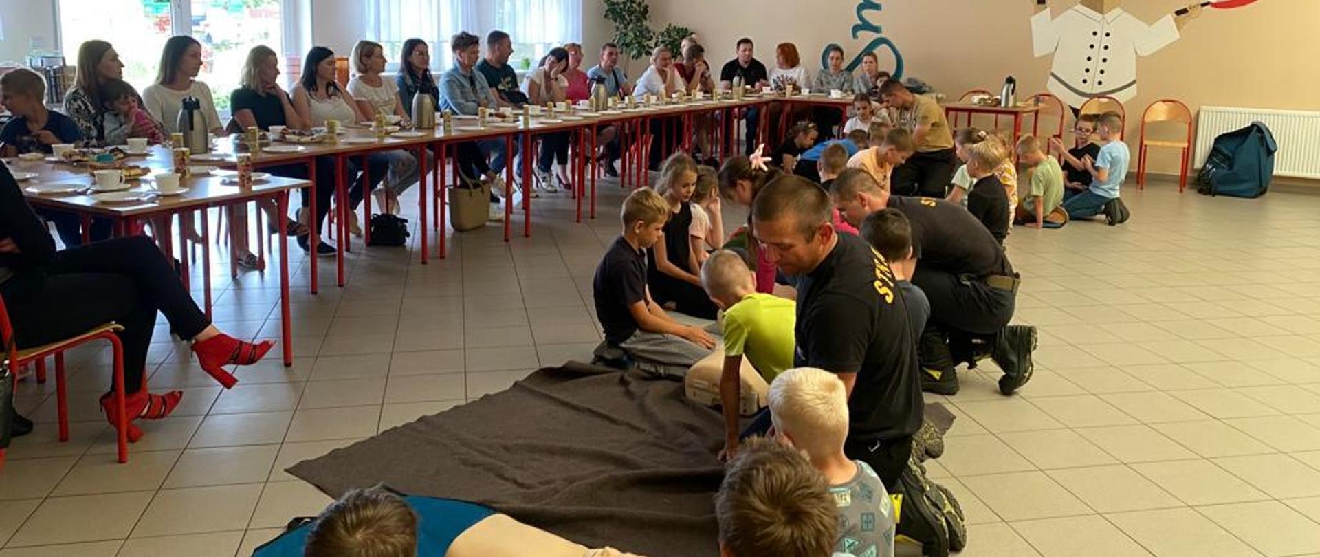 Zdjęcie przedstawia strażaków nadzorujących dzieci podczas wykonywania masażu serca. Na drugim planie znajdują się ich rodzice i opiekunowie siedzący przy stolikach szkolnych.