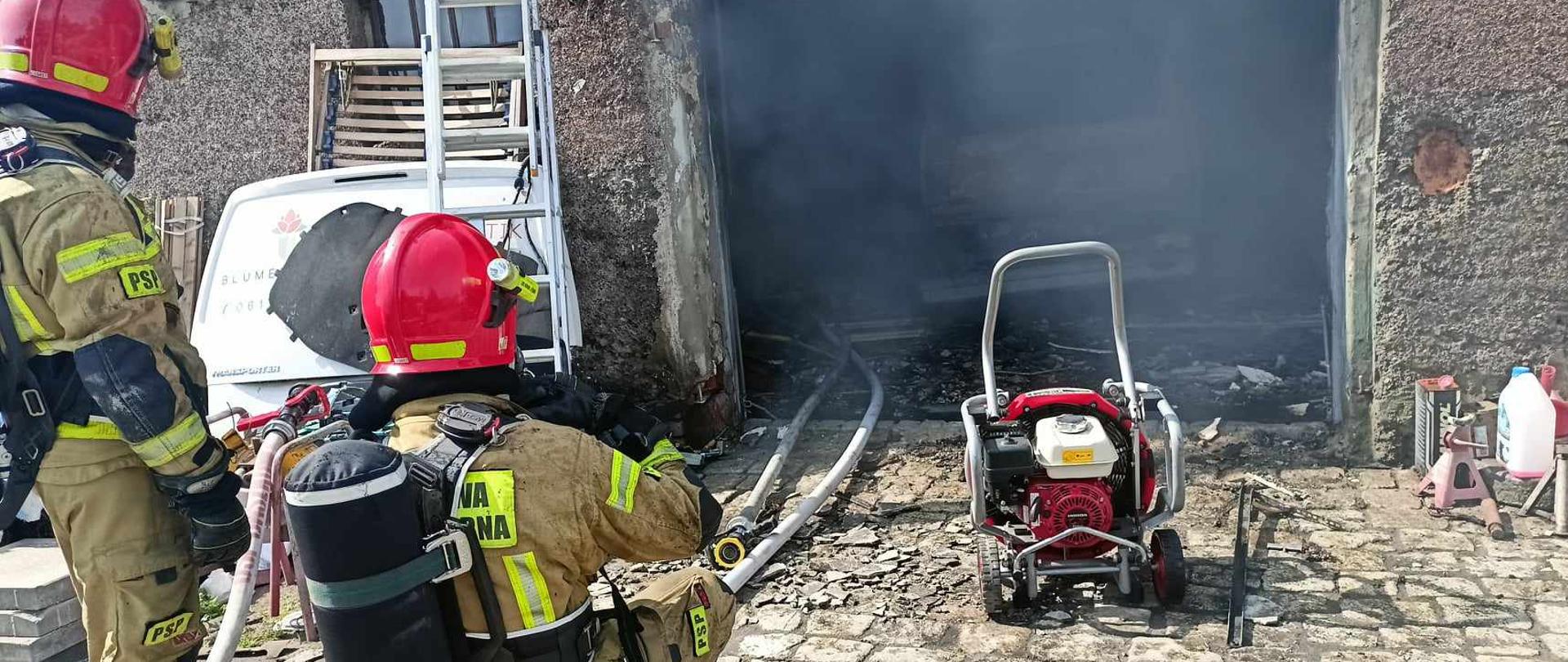 Zdjęcie przedstawia strażaków, którzy prowadzą pomiar temperatury wewnątrz palącego się obiektu. Przed bramą garażową wentylator osiowy, wewnątrz samochód, który uległ spaleniu.