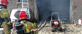 Zdjęcie przedstawia strażaków, którzy prowadzą pomiar temperatury wewnątrz palącego się obiektu. Przed bramą garażową wentylator osiowy, wewnątrz samochód, który uległ spaleniu.