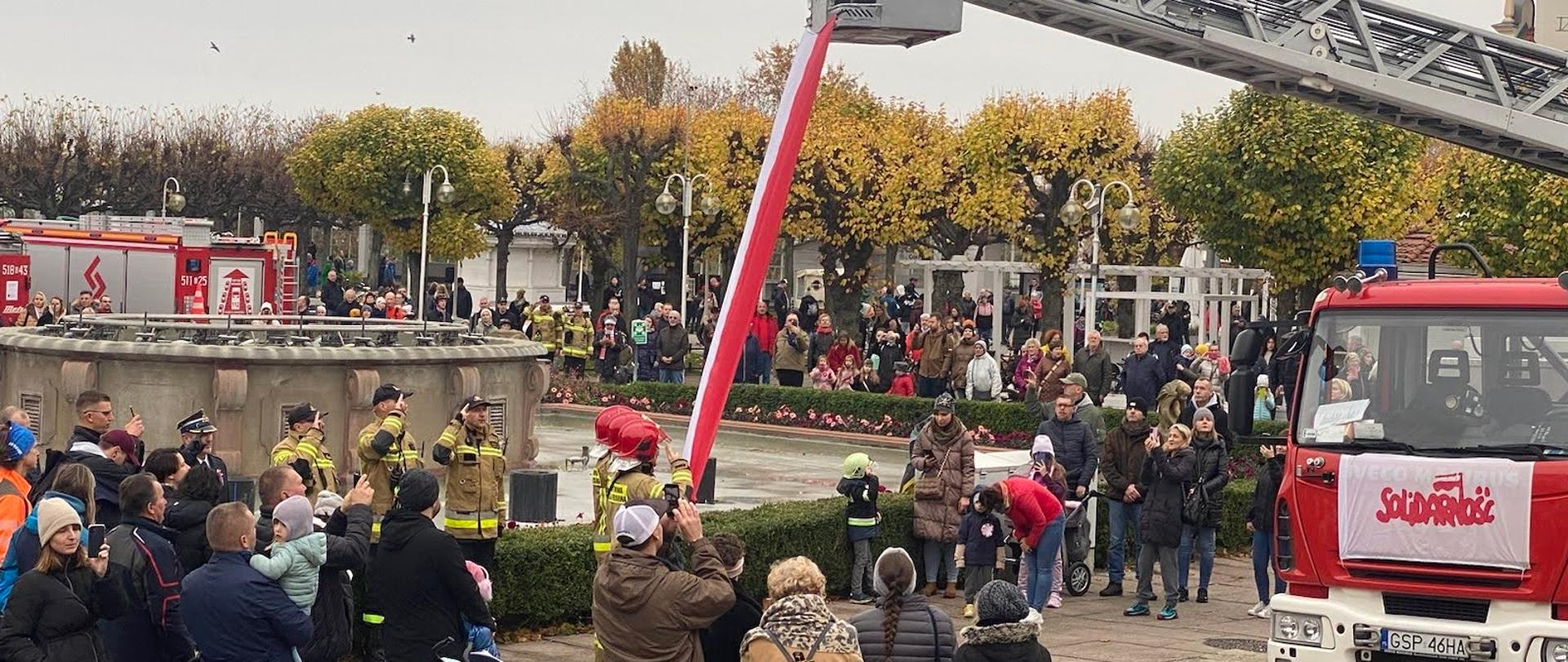 Strażacy oddają honor podczas wciągnięcia flagi obok stoją ludzie.