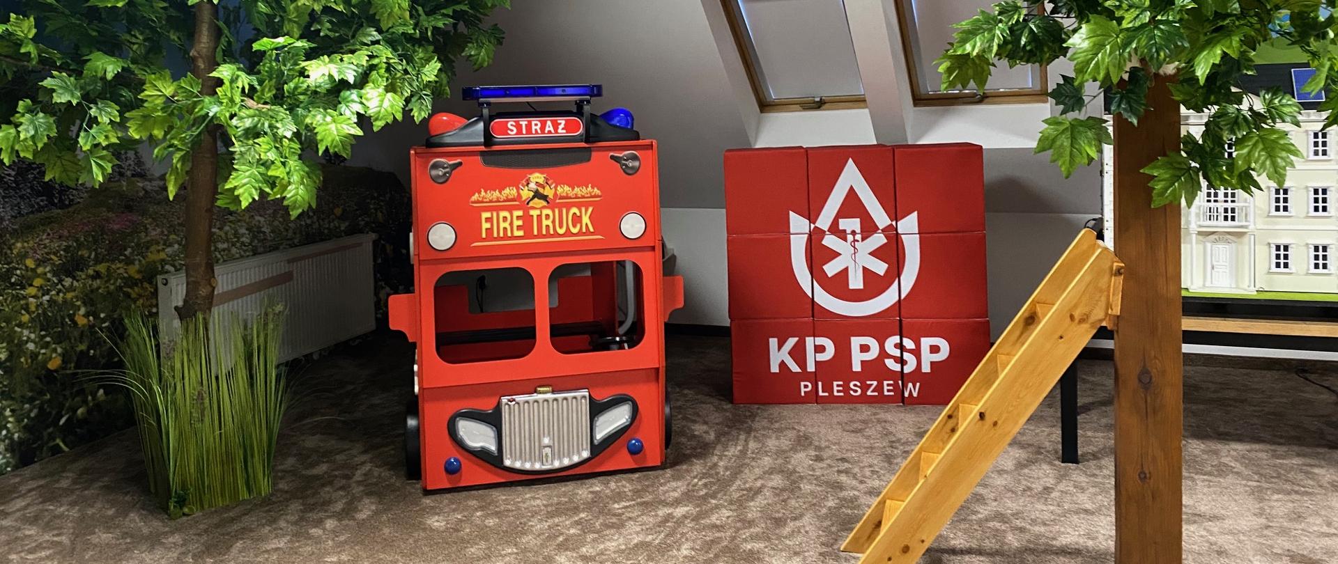 Imitacja samochodu strażackiego dla dzieci. Sztuczne drzewka oraz drabinka oparta o jedno z nich.