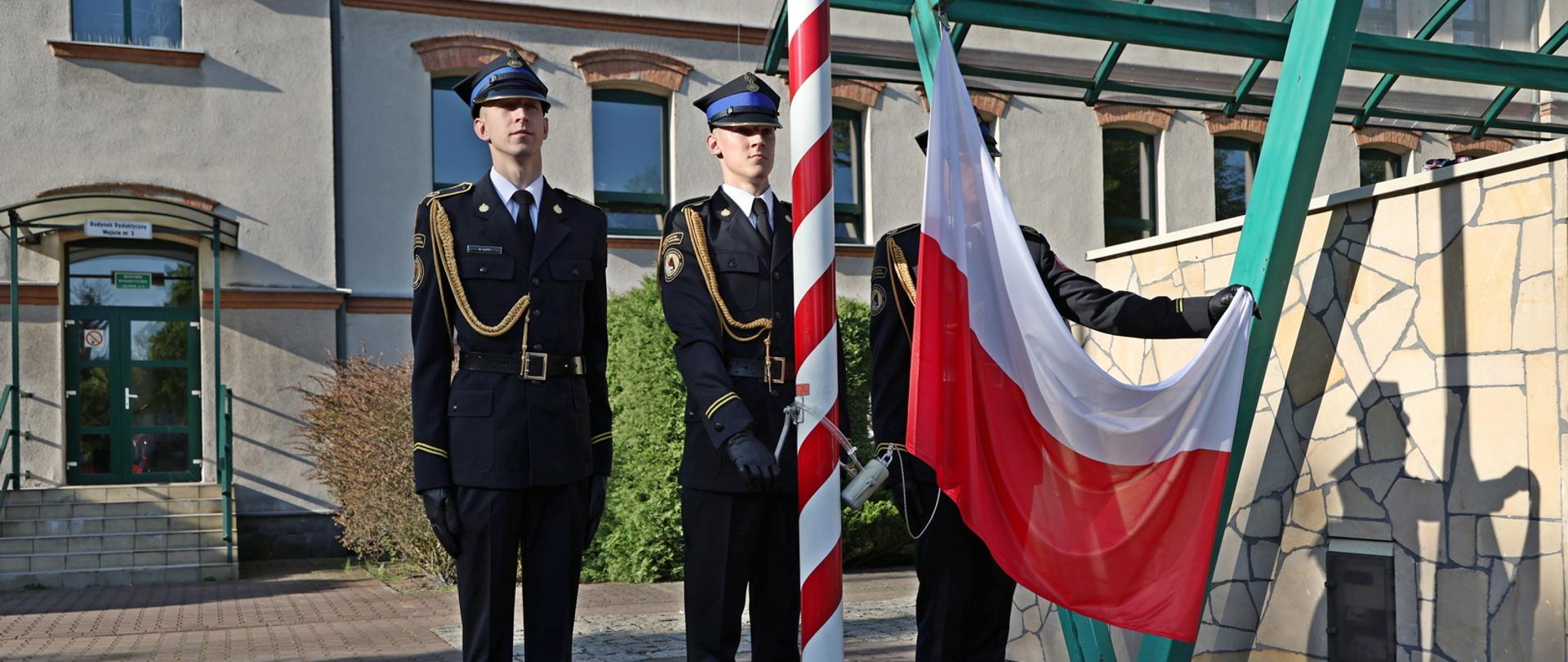 Kadeci pocztu flagowego podczas wciągania flagi Rzeczypospolitej Polskiej na maszt, w tle budynek dydaktyczny Centralnej Szkoły PSP