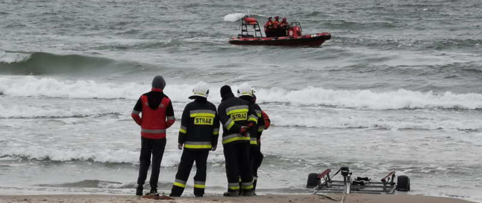 Brzeg morza, na plaży stoją strażacy, obok przyczepa do przewozu łodzi, w oddali na morzu ponton z ratownikami