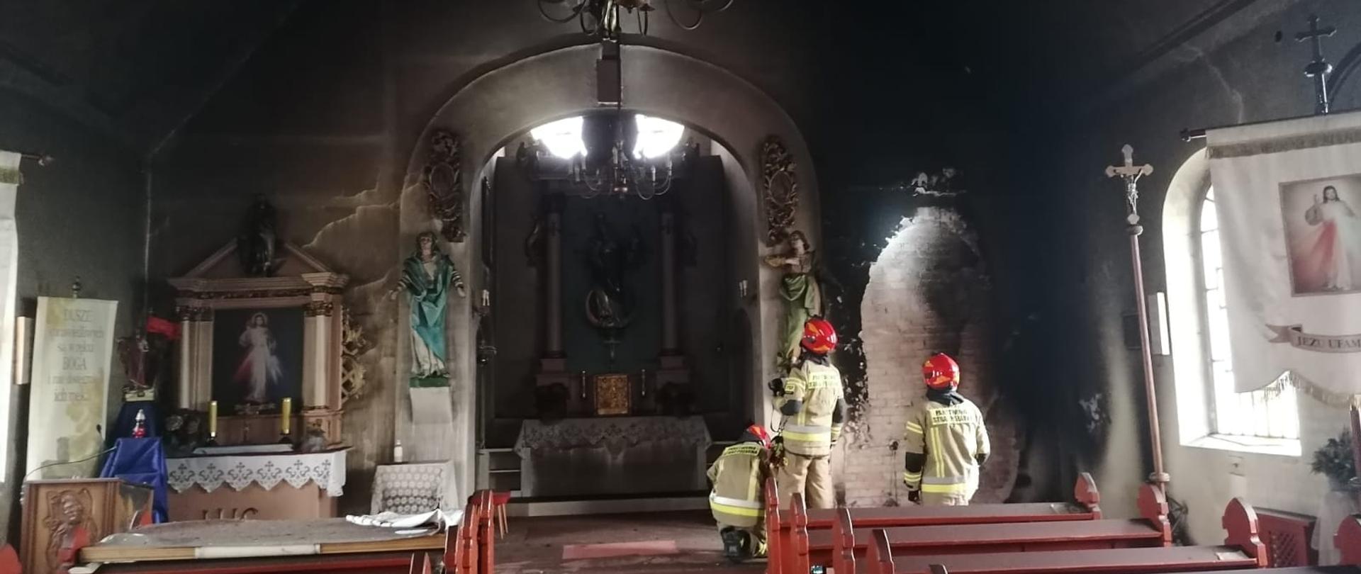 Wnętrze kościoła po pożarze. Czarne ściany i sufit świątyni. Wewnątrz trzech strażaków w jasnych mundurach i czerwonych hełmach.