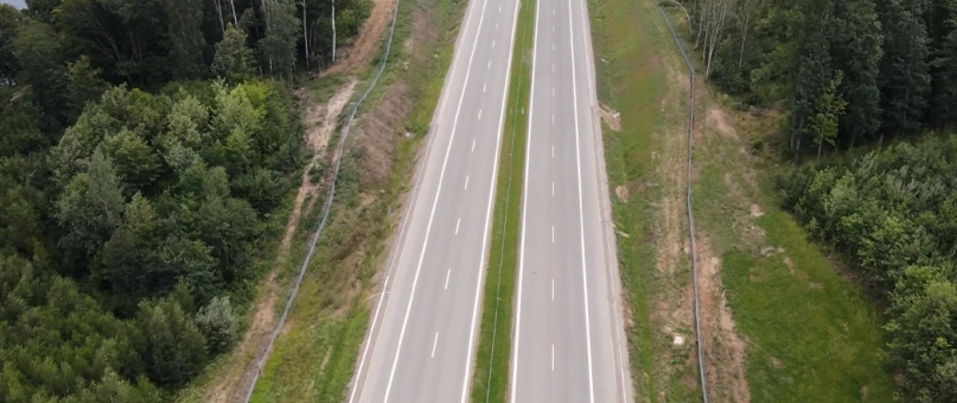 Dwupasmowa droga z pasem zieleni pośrodku, biegnąca przez zalesiony teren, widziana z góry.