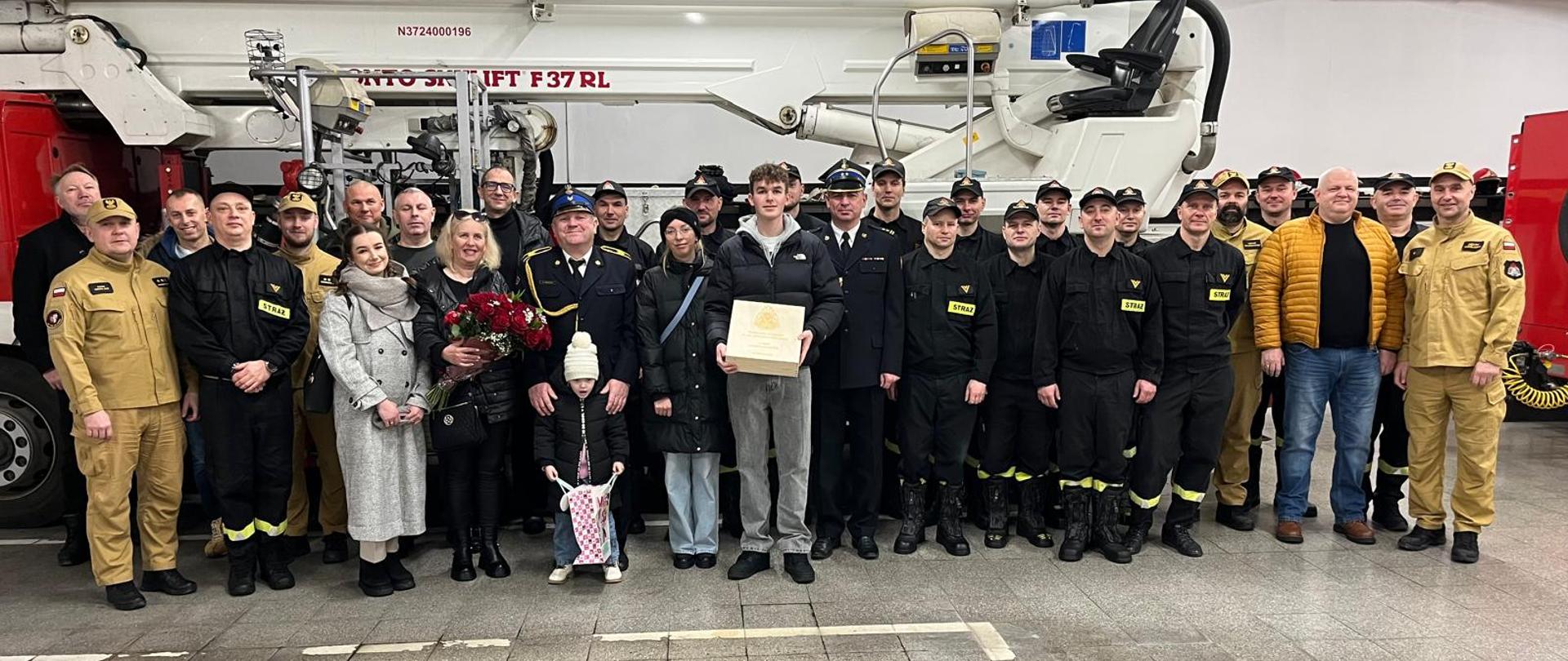 Grupa strażaków w umundurowaniu roboczym i galowym oraz osób towarzyszących pozuje do wspólnego zdjęcia w remizie strażackiej. W tle widoczny jest wóz strażacki oraz wyposażenie hali.