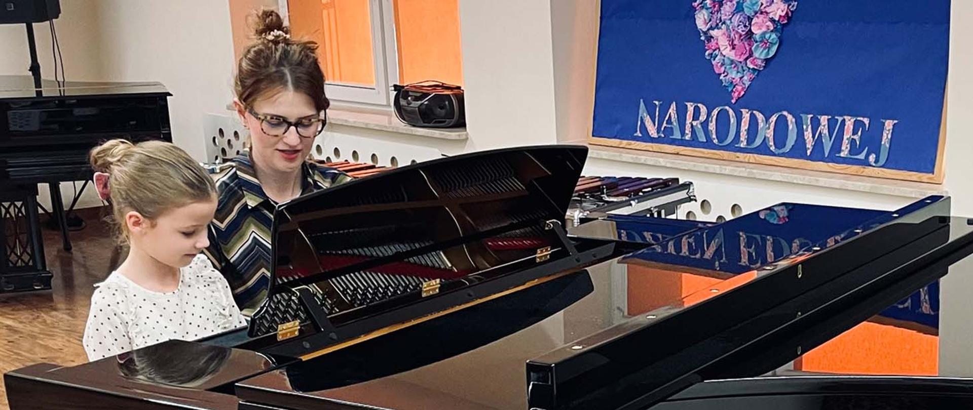Na zdjęciu nauczycielka i uczennica w auli grają w duecie na fortepianie, widoczne ich twarze i fragment fortepianu bez klawiatury. W tle ściana z fragmentem planszy na której widnieje napis o treści narodowej.