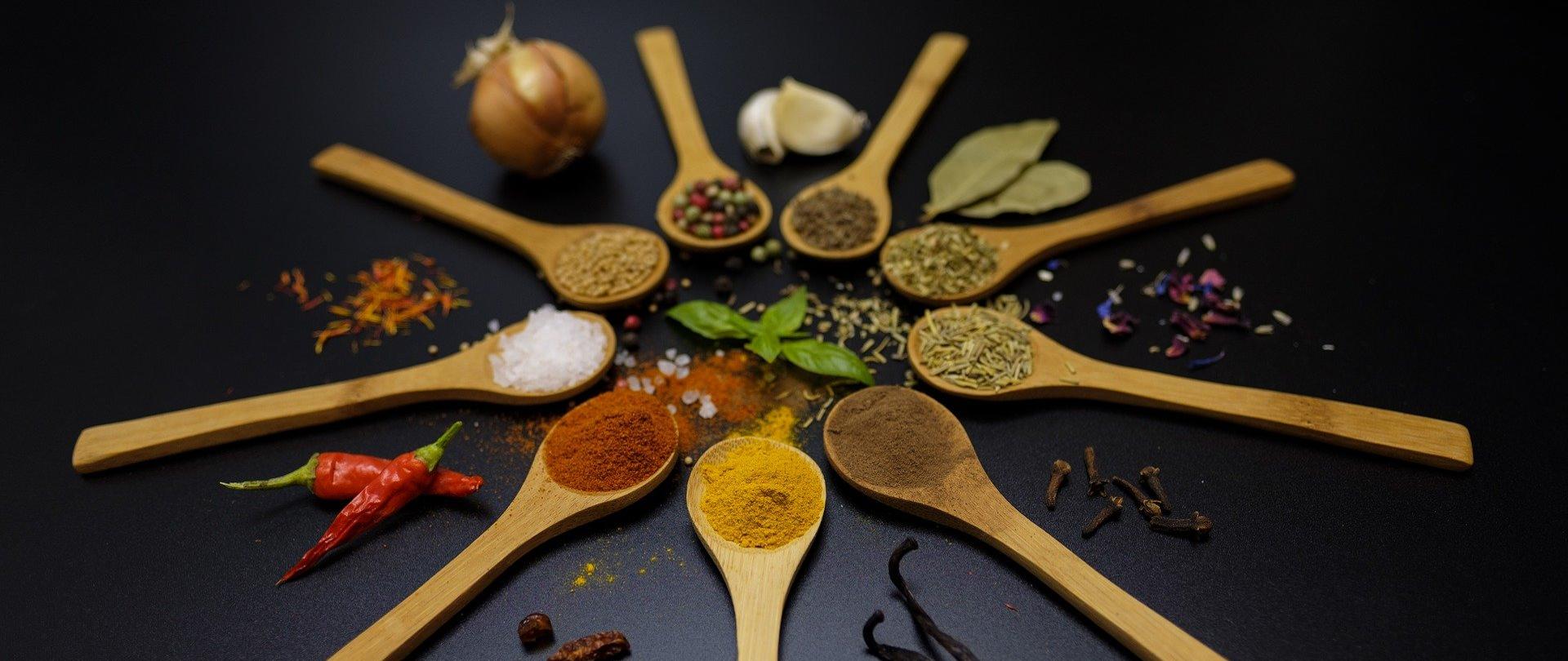 Na drewnianych łyżkach ułożonych w okrąg znajdują się różne przyprawy: papryka, kurkuma, pieprz, kminek, kolorowy pieprz, gruboziarnista sól.