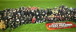 Grupowe zdjęcie uczestników nocnych zawodów strażackich na oświetlonym boisku. Strażacy pozują z pucharami i dyplomami, a przed nimi leży baner „Ochotnicza Straż Pożarna w Bluszczowie”.