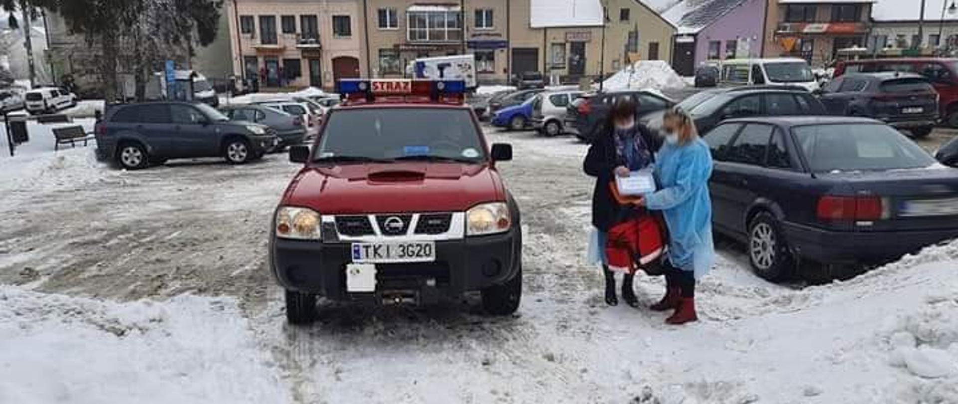 Zdjęcie przedstawia samochód operacyjny Ochotniczej Straży Pożarnej w Łagowie stojący na parkingu przed ośrodkiem zdrowia. Po prawej stronie znajdują się dwie kobiety. W tle widnieje rynek w Łagowie.