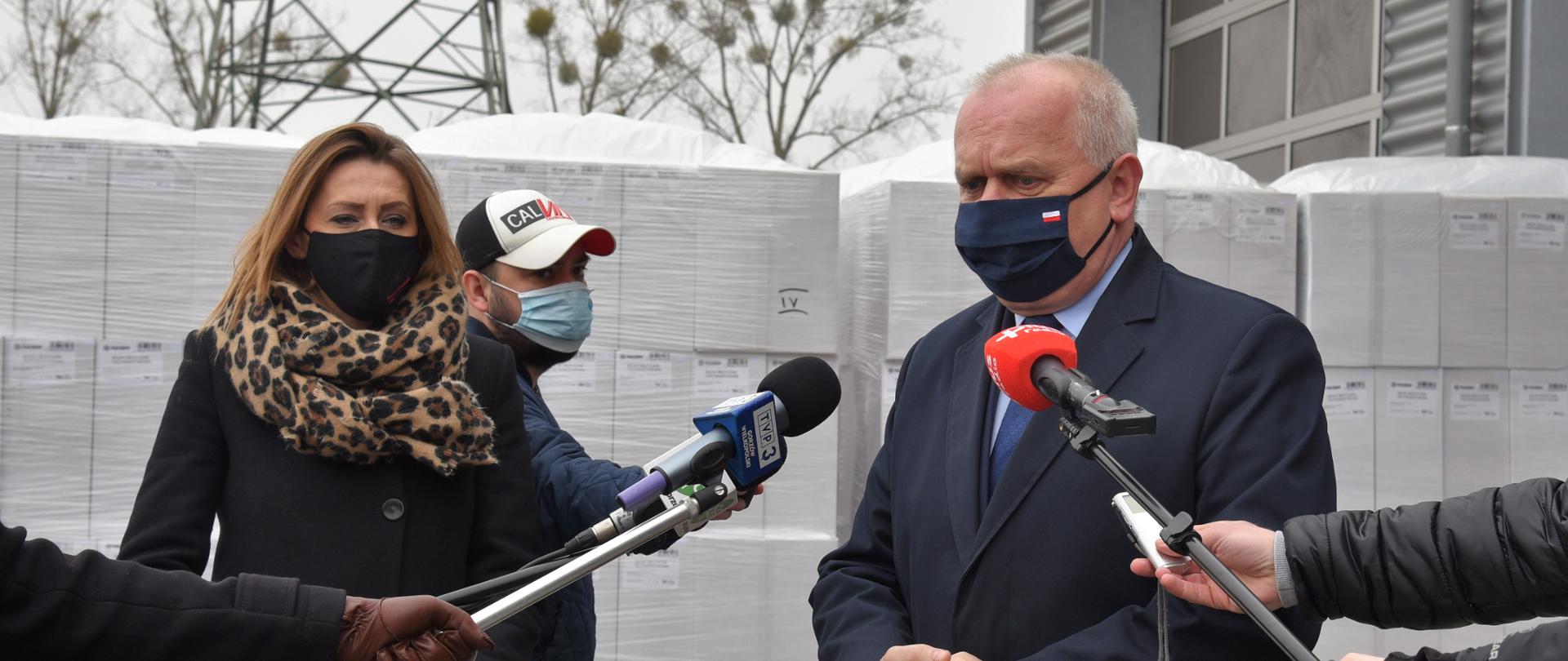Wojewoda ze starostą gorzowską w czasie briefingu prasowego, w tle palety z kartonami z maseczkami, po bokach ręce dziennikarzy z wyciągniętymi mikrofonami 