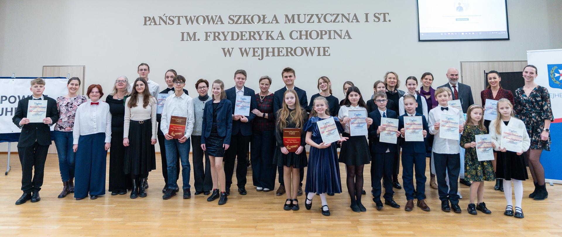 Na sali koncertowej pozują na stojąco wraz z dyplomami uczestnicy konkursu wraz z nauczycielami oraz jurorami.