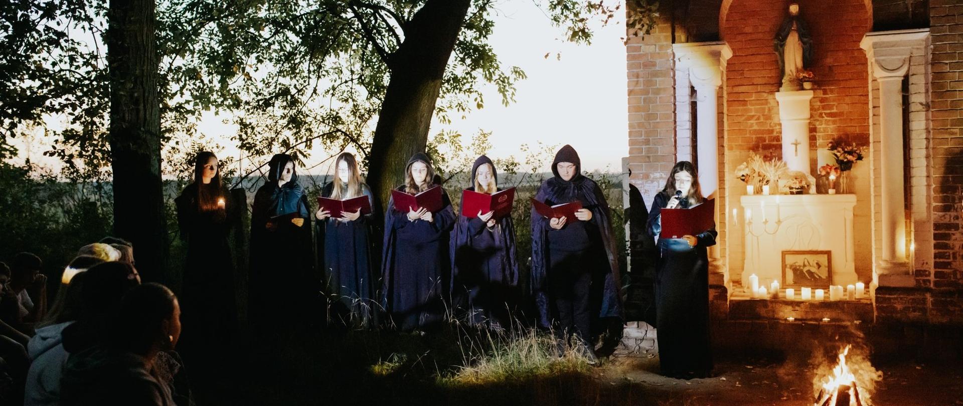Zdjęcie przedstawia Narodowe Czytanie „Dziadów” Adama Mickiewicza. Przed oświetloną kapliczką stoi grupa uczestników ubranych w ciemne, długie peleryny. Osoby trzymają otwarte czerwone księgi i odczytują fragmenty utworu, a jedna z nich korzysta z mikrofonu. Wokół kapliczki ustawiono świece, a na pierwszym planie płonie ognisko. Po lewej stronie widoczna jest zgromadzona publiczność.
